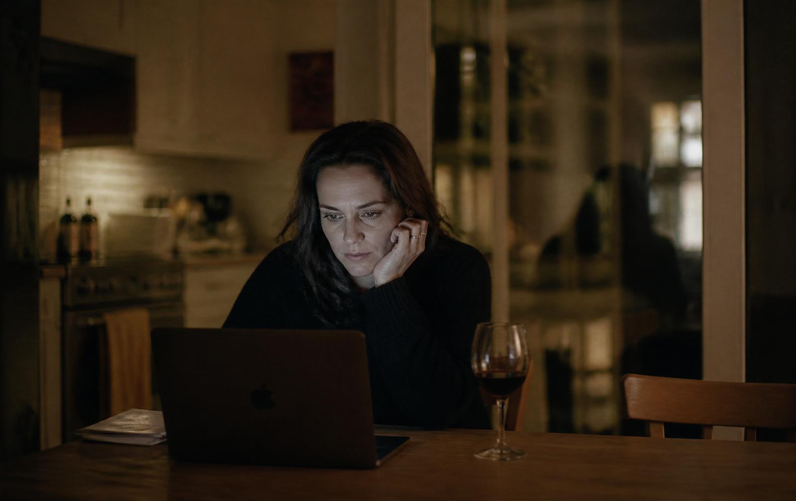 A woman sits alone at her kitchen table late at night, illuminated by her laptop screen.