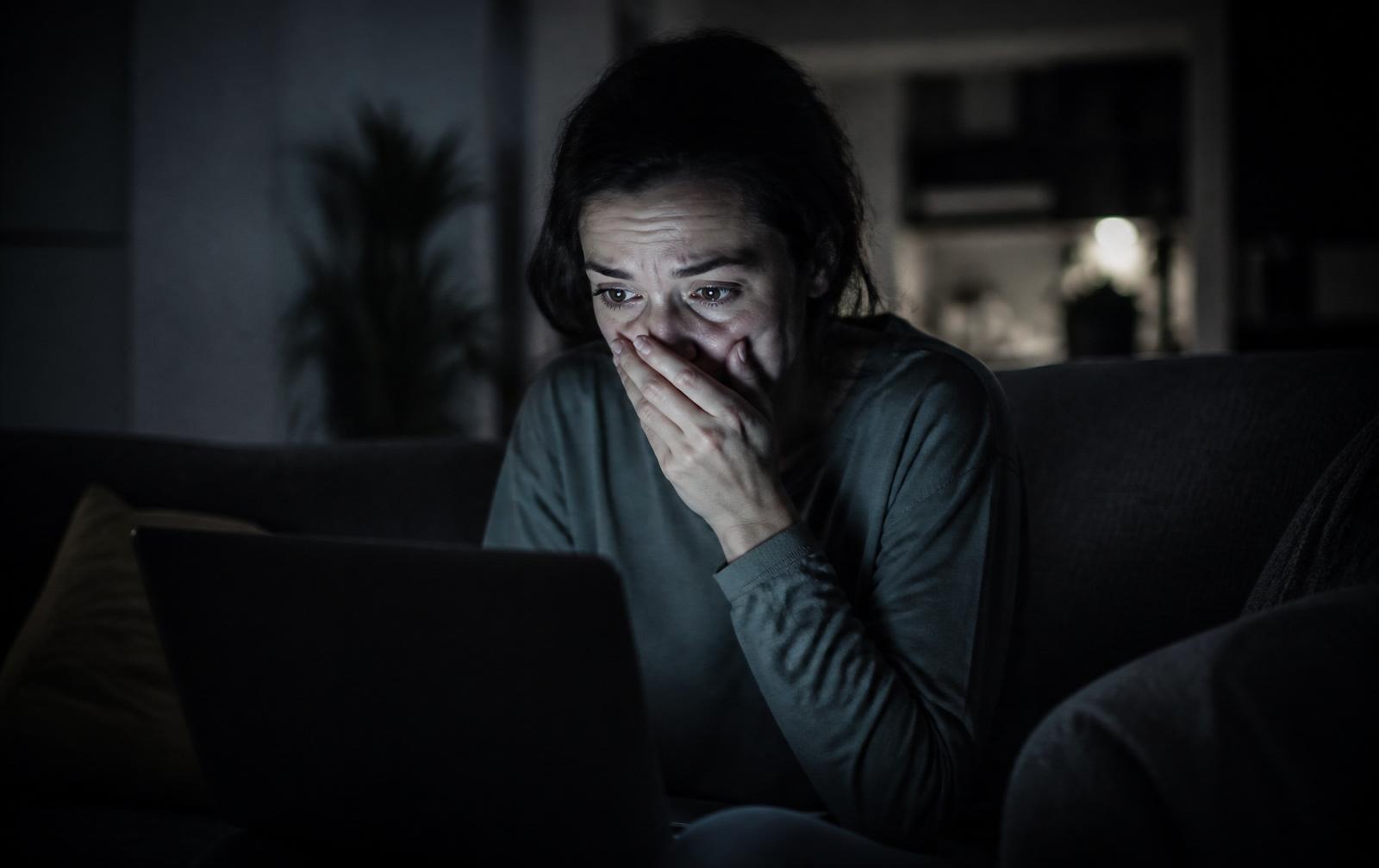 A woman sits in a dark living room lit only by her laptop screen, one hand covering her mouth.